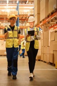 Two workers walking through large warehouse with pallets of boxes.