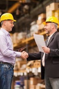 Two Businessmen Shaking Hands and Wearing Safety Hats while Standing in Front of their Company's Products