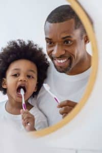 Father looking in mirror while teaching young child how to brush their teeth.