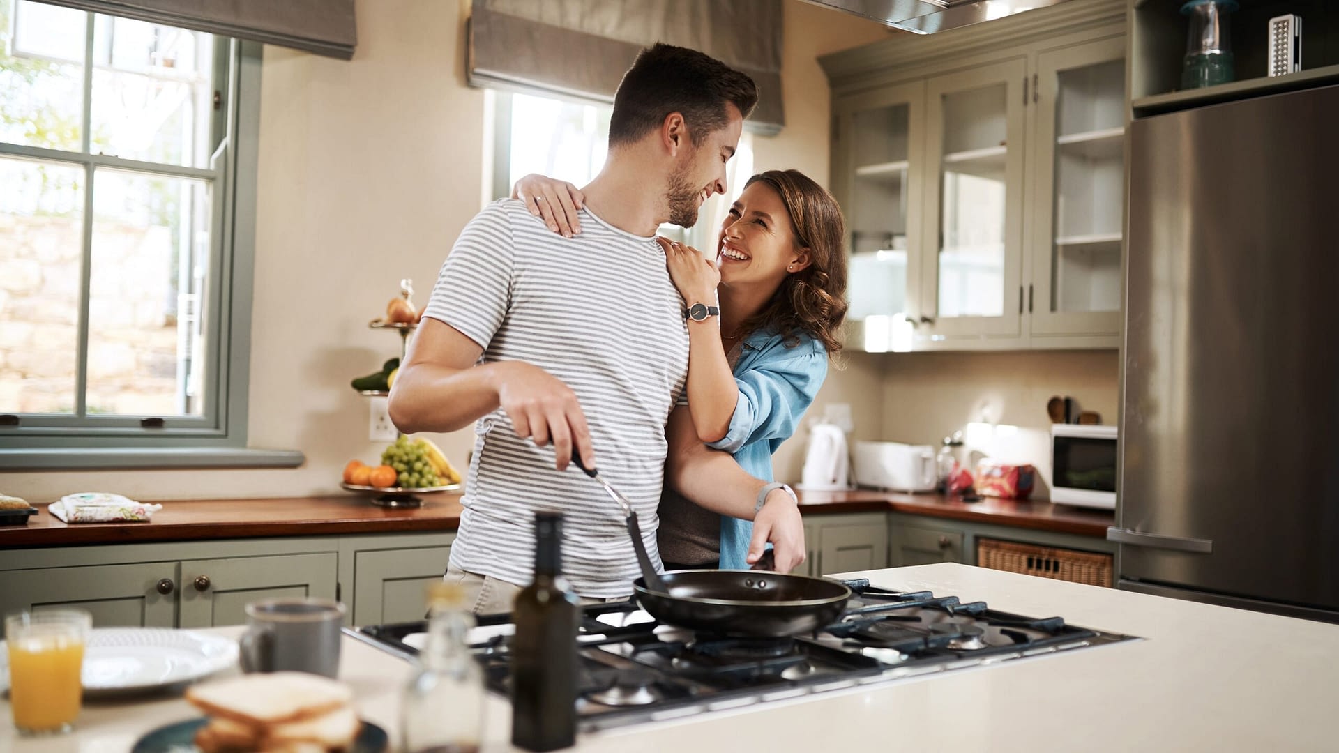 Young woman embracing her partner while he cooks breakfast in the kitchen.
