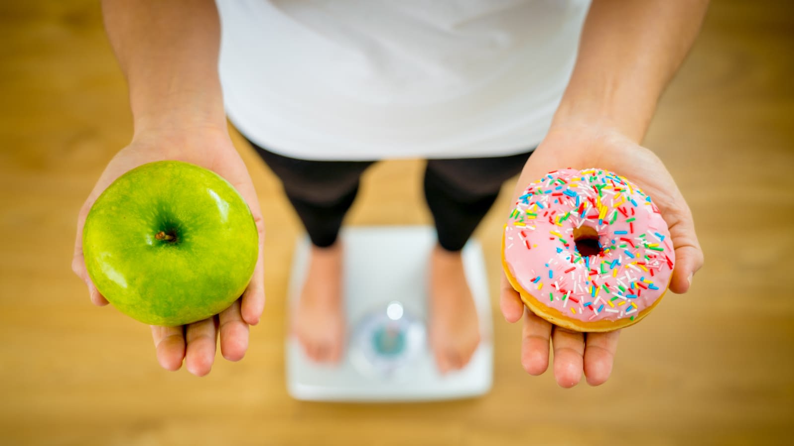 Close up of woman on scale holding on hands apple and doughnut making choice between healthy unhealthy food dessert while measuring body weight in Nutrition Health care Diet and temptation concept. Person on scale with food