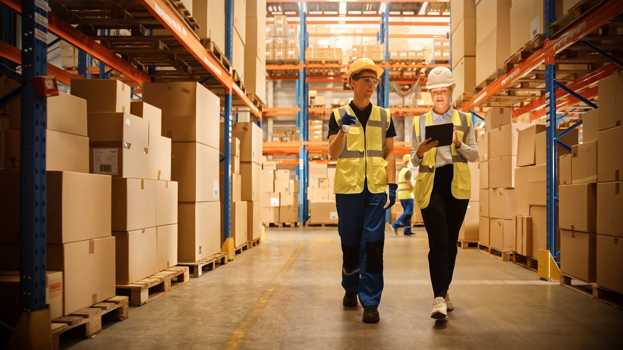 Two workers walking through large warehouse with pallets of boxes.