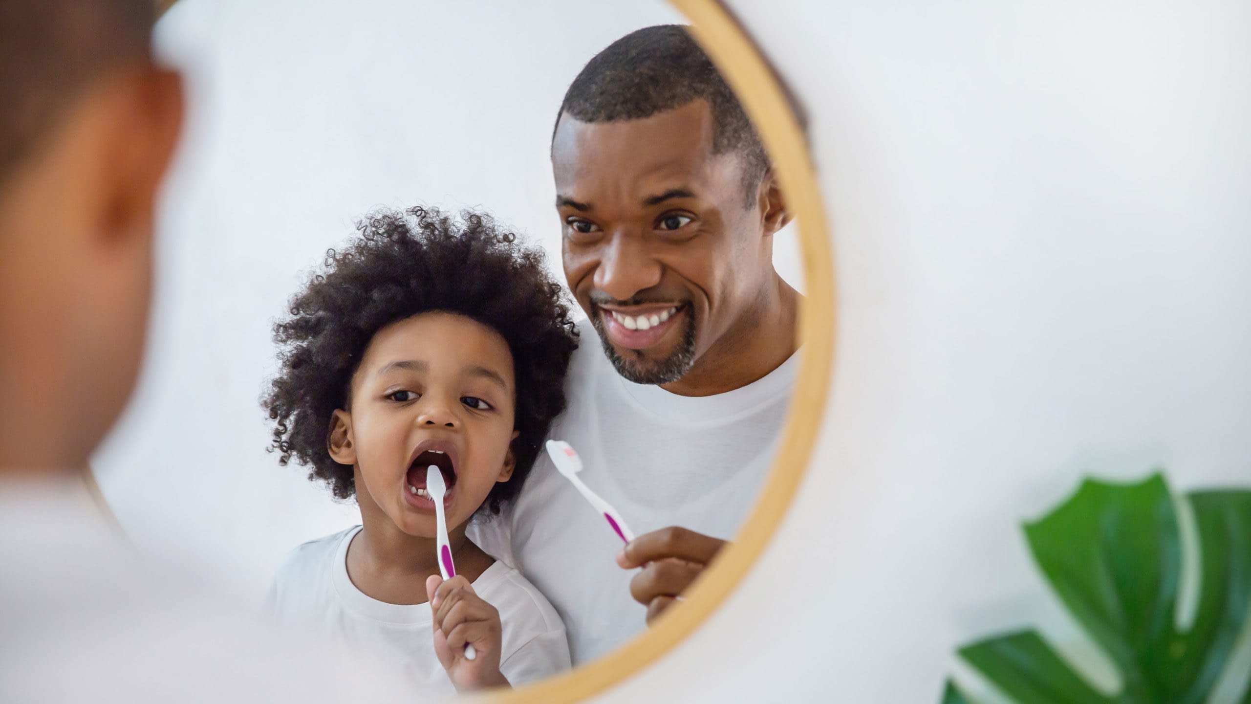 Father looking in mirror while teaching young child how to brush their teeth.