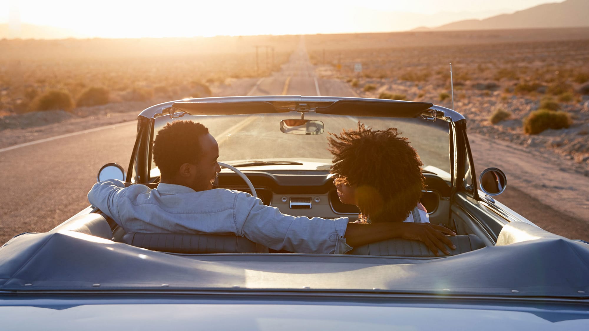 Couple in a convertible car with the top down driving at sunset.