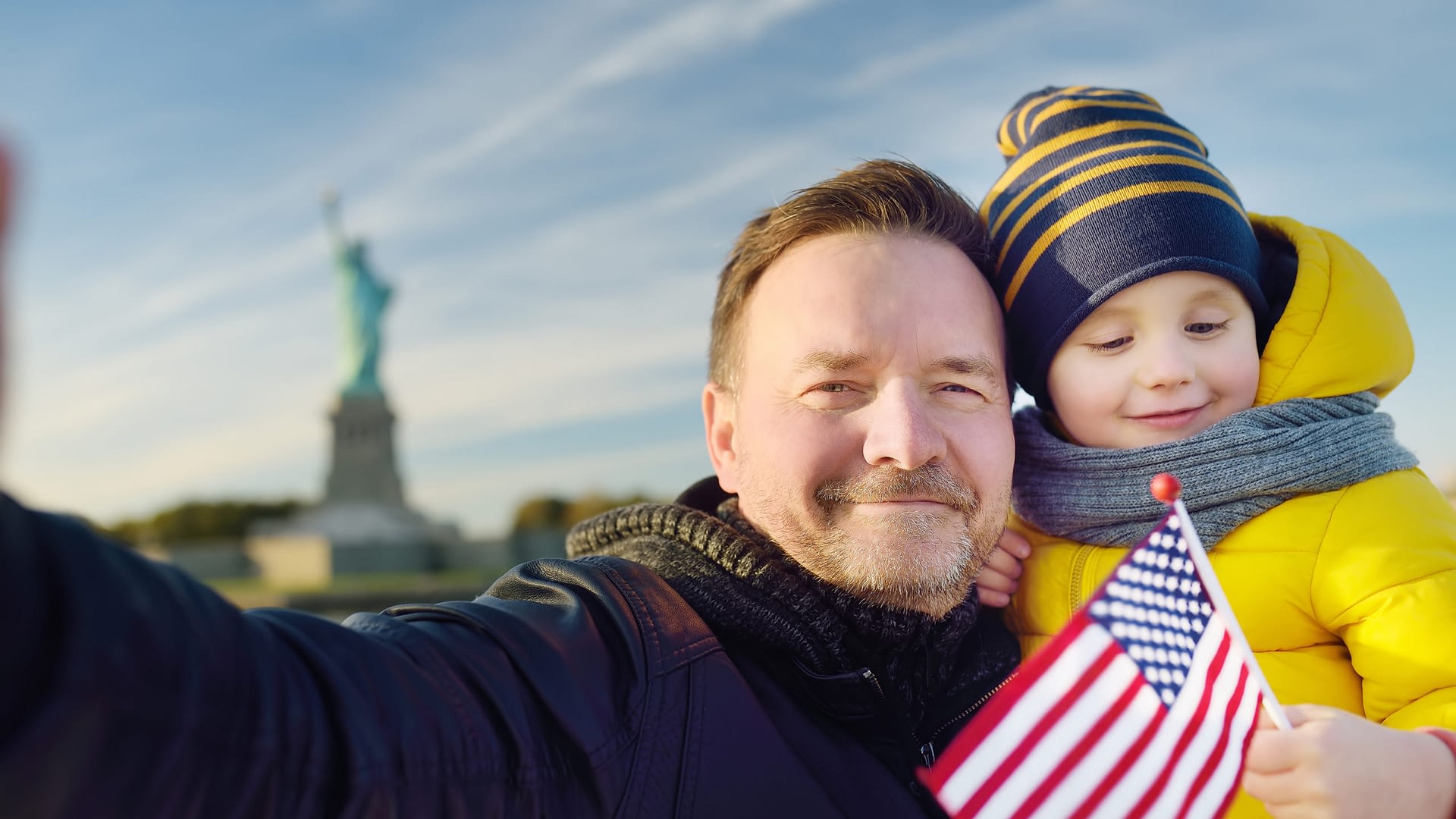 Father and his little son holding the American flag on the background of the statue of liberty