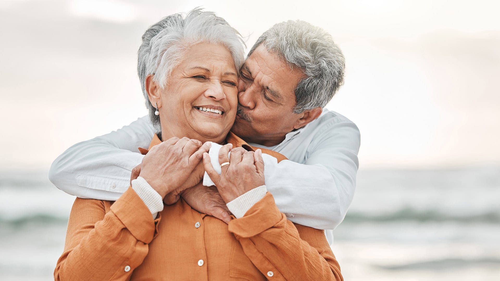Husband kissing wife on cheek with beach in the background.