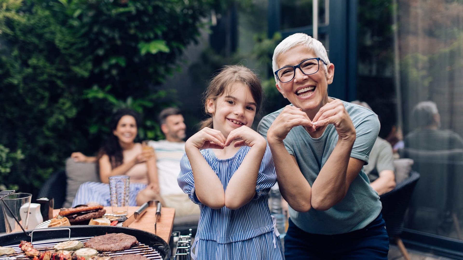 Little girl helping her grandma preparing barbecue for the whole family at the backyard and showing hearts.