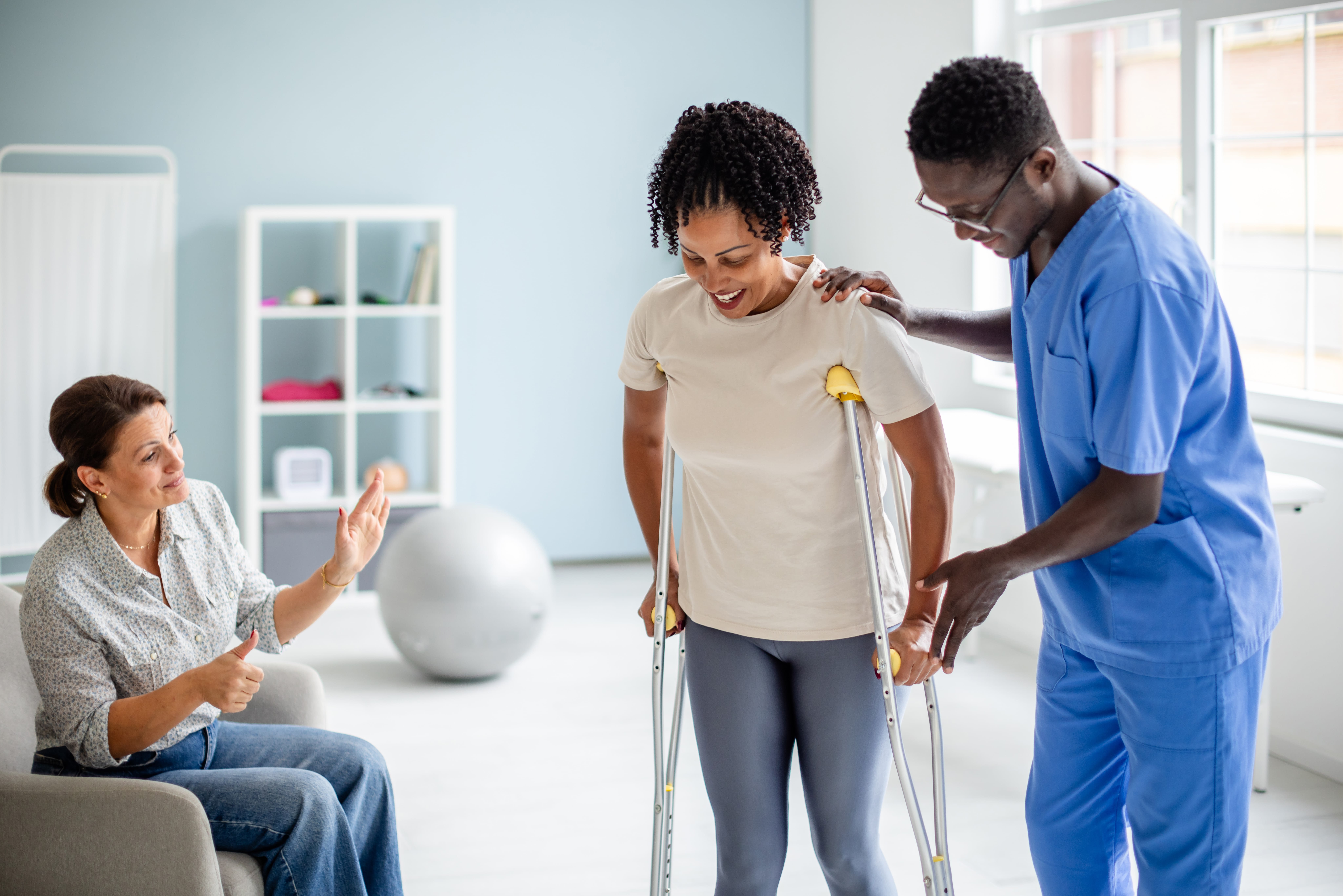 A physical therapist supports a patient using crutches as another person watches and encourages with a thumbs-up gesture, emphasizing teamwork and rehabilitation progress.