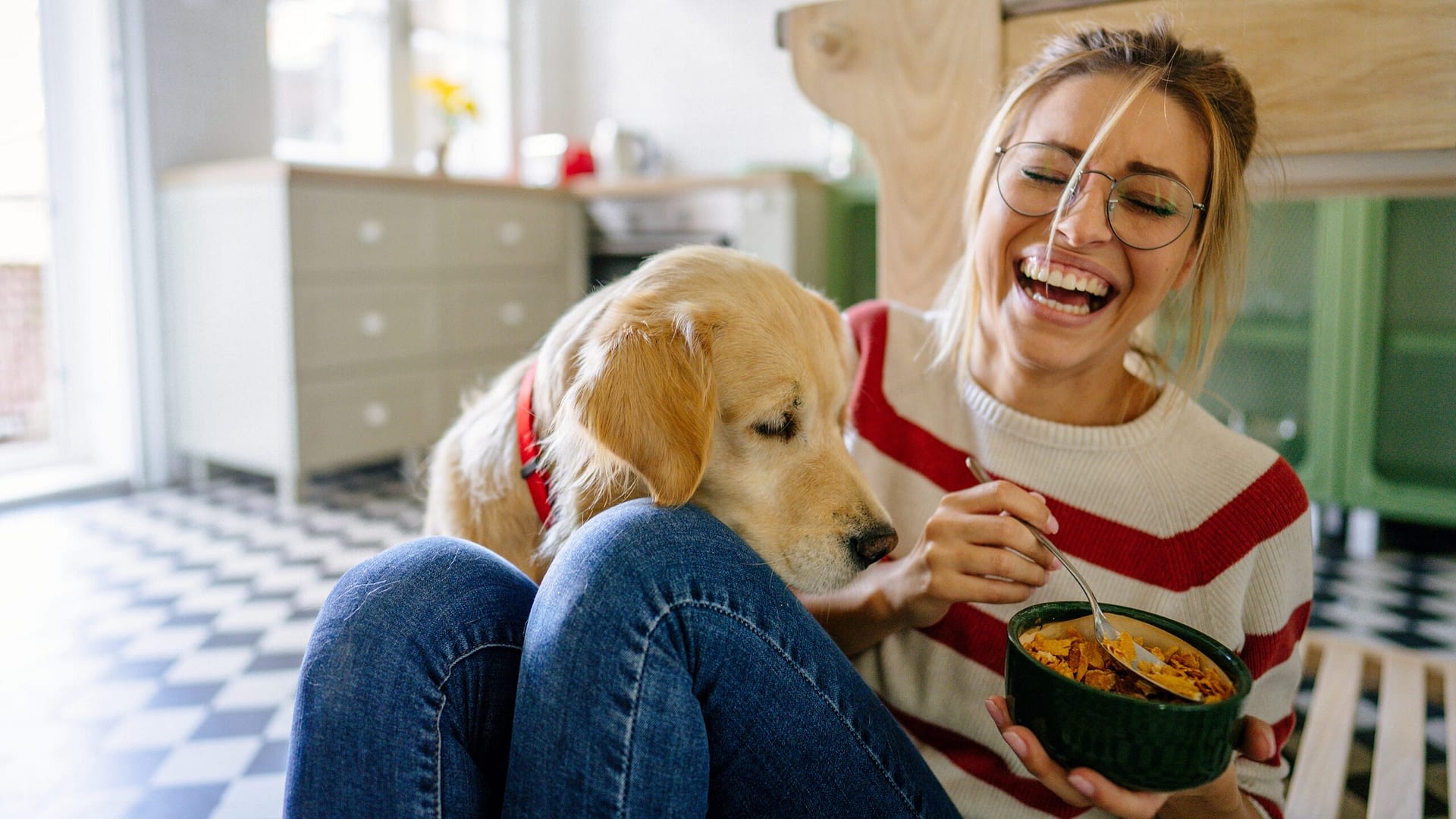 Young adult woman laughing and sitting on the floor with her dog while holding a bowl of food.