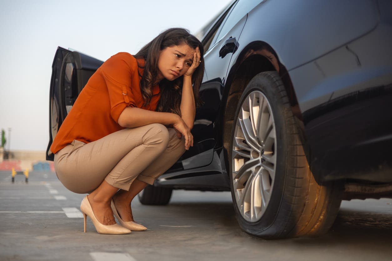 Don’t Neglect Your Car’s Tires They Are Lifesavers Portrait of a despair woman crouching next to her car with flat tire, problems on the road, deflated tire.