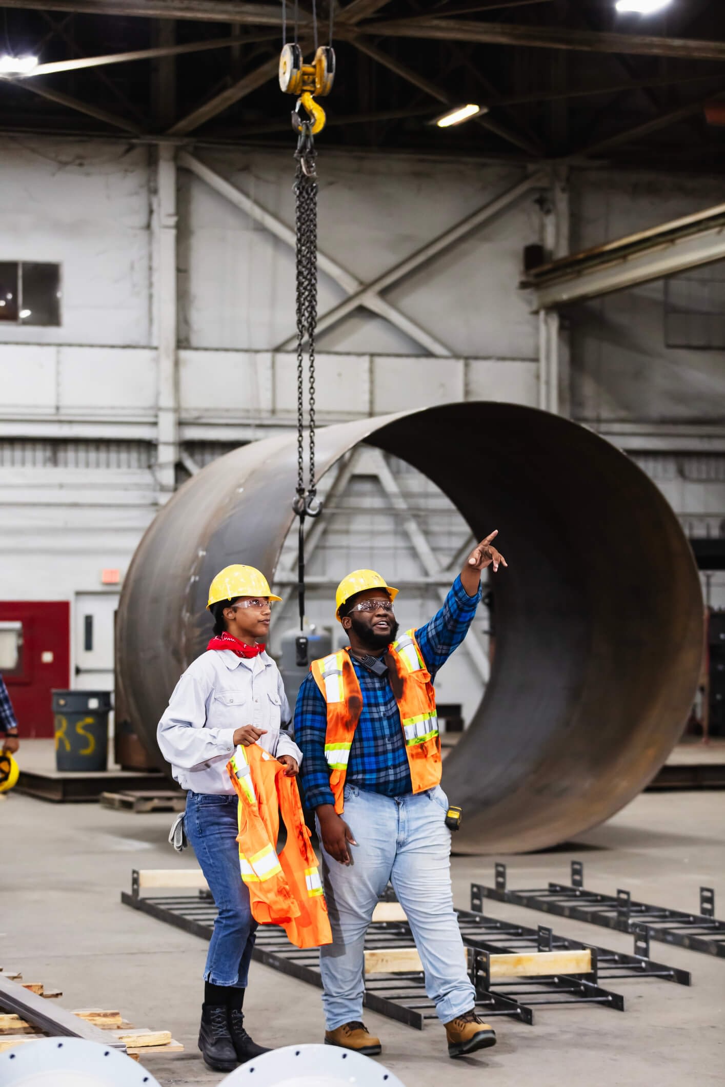 Two young African-American workers, a man and a woman, walking in a steel fabrication factory, conversing. The man is looking up and pointing at something.