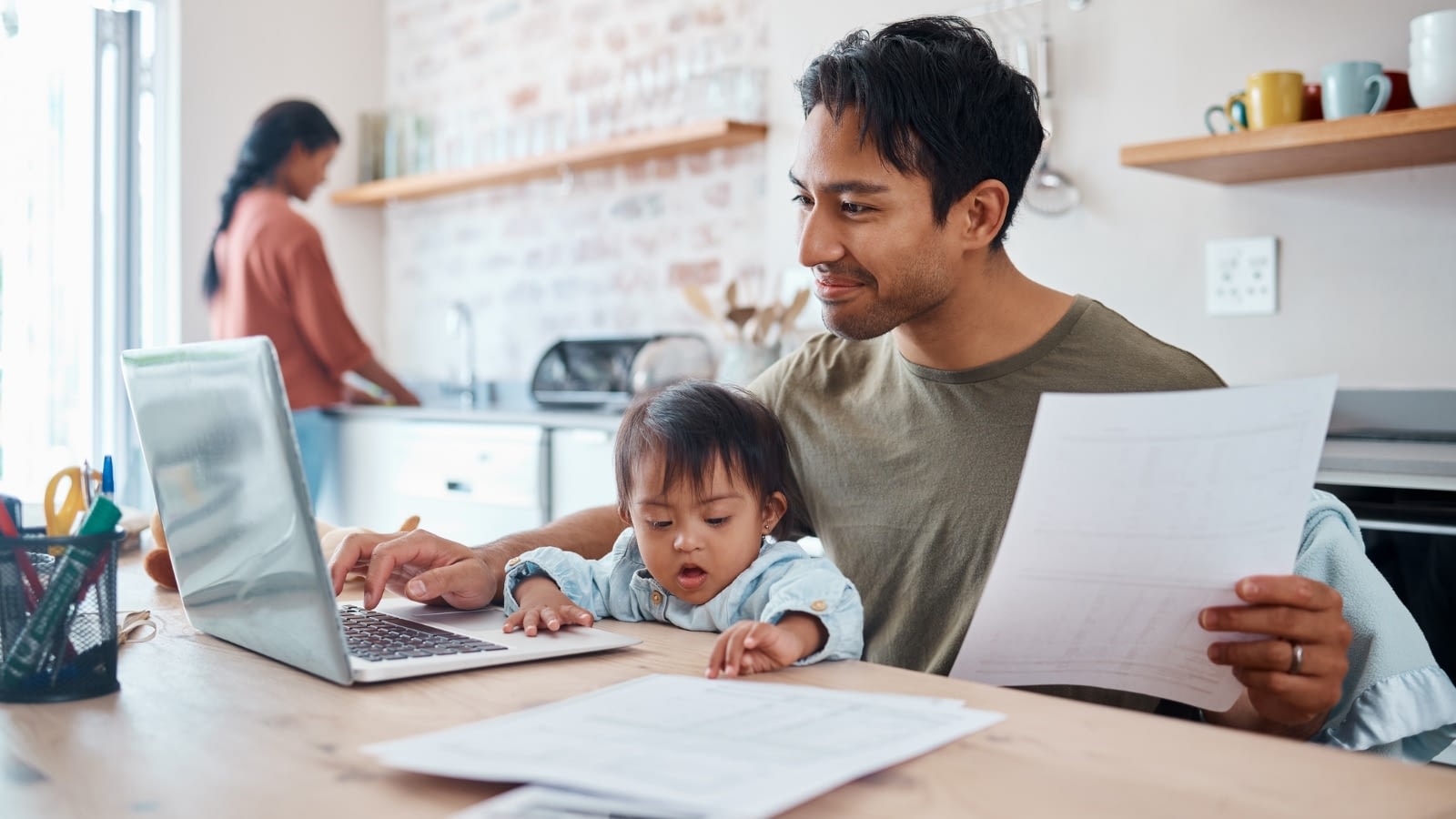 Young man with toddler in lap and wife in background researching on. his laptop.