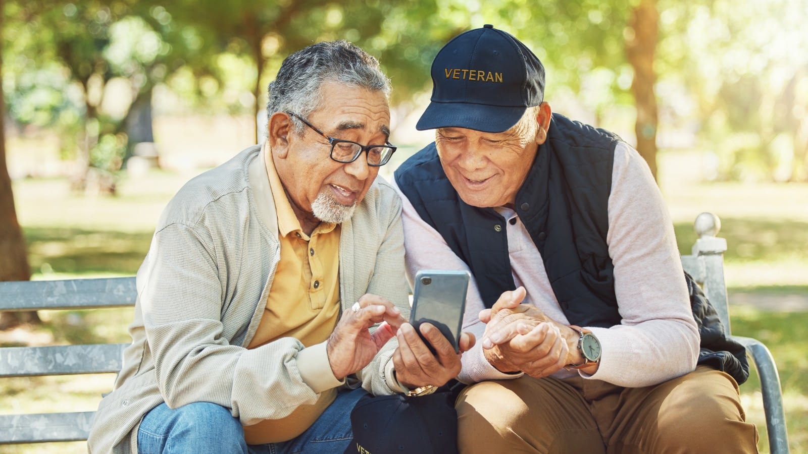 two elderly veterans sitting on park bench looking at something on cell phone.