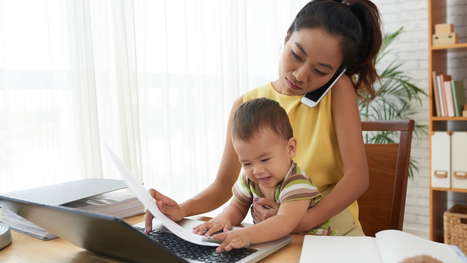 young Asian mom working from home while holding baby