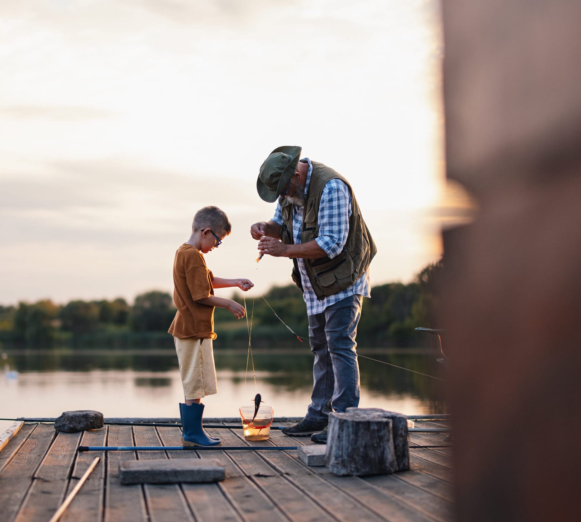 An older man teaches a young boy how to fish on a wooden dock by a serene lake. The scene captures the beauty of nature and the warmth of family connection.