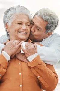 Husband kissing wife on cheek with beach in the background.