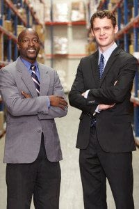 Two Businessmen Stand Beside Each Other in Front of Their Company's Products With Their Arms Crossed