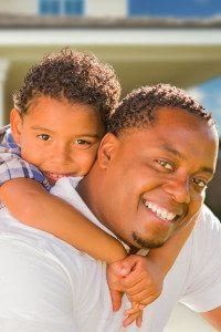 Adult Smiling Male Carries a Smiling Young Boy on his Back Standing in Front of a Home