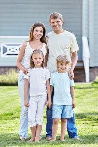 Husband, Wife, Daughter, and Son Stand Smiling at the Camera in Front of their Home