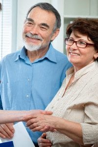 Both a Male and Female Employee Stand and Smile at Another Individual while the Female Shakes a Hand and the Male Holds Tax Documents