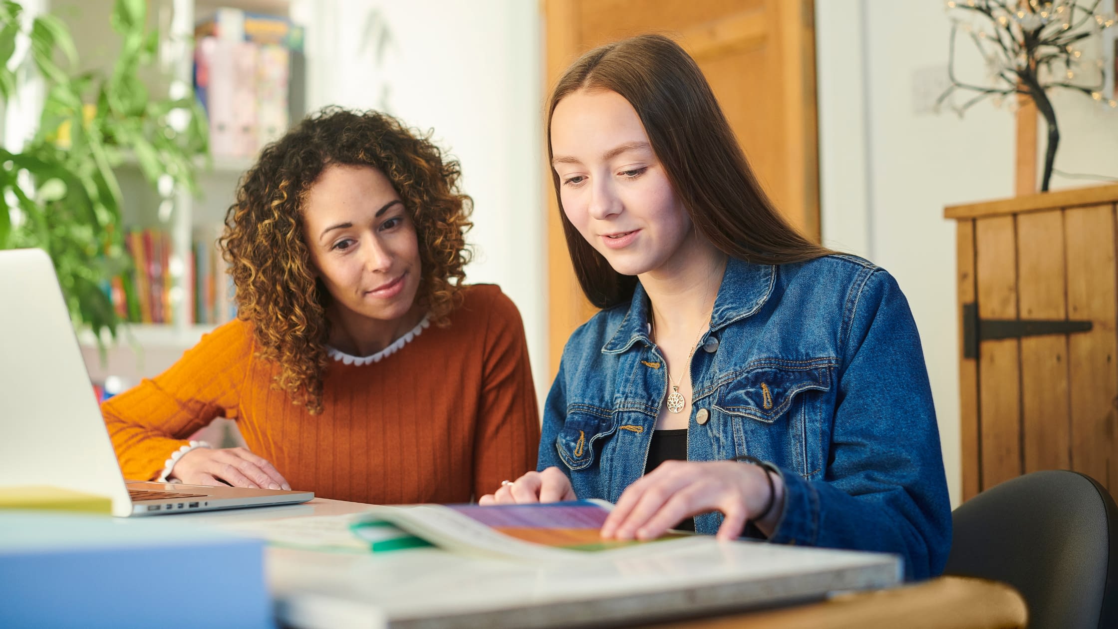Untitled design Woman tutoring girl at table, surrounded by books and computer.