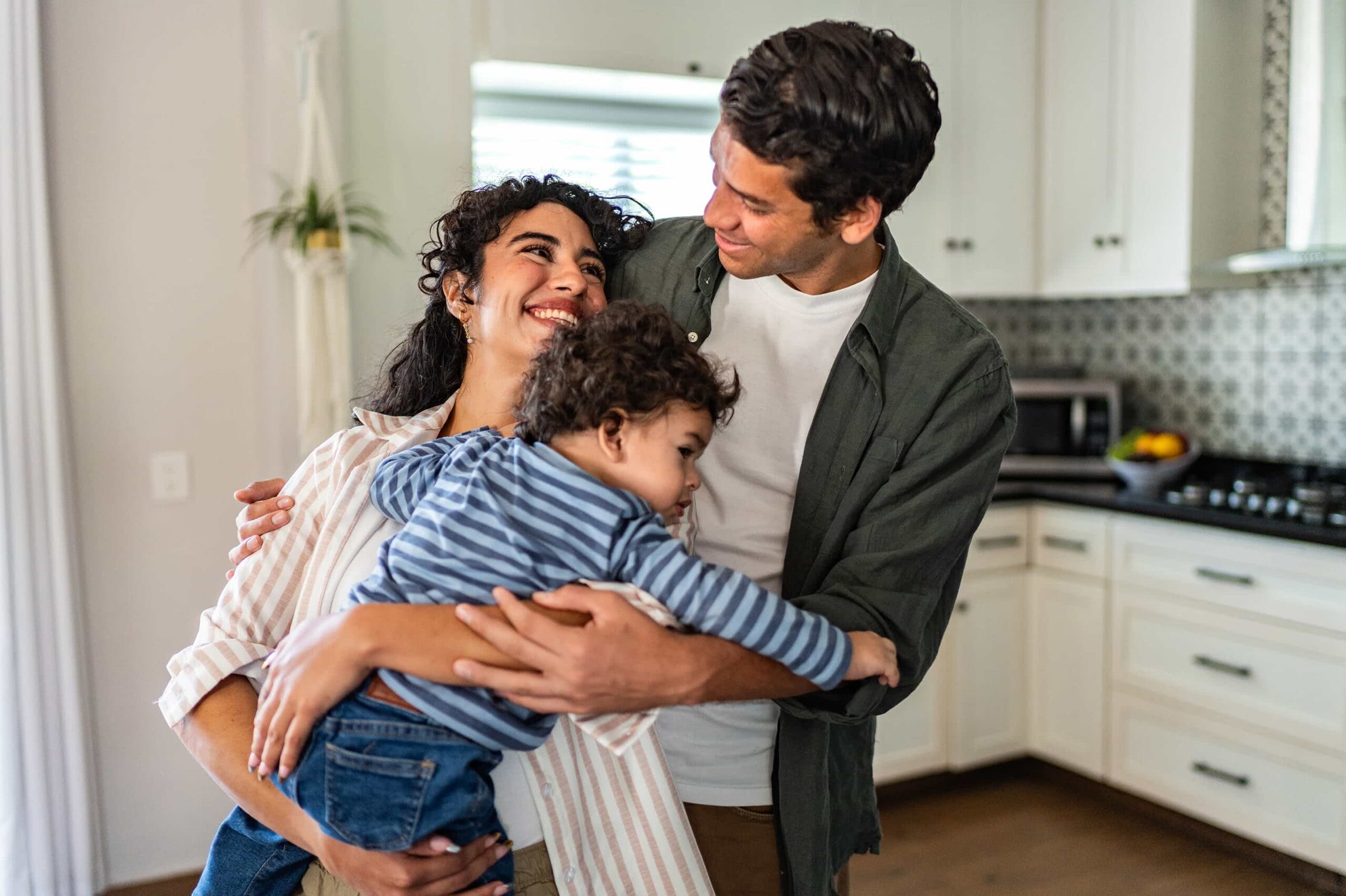 A happy mother and father smiling at each other while embracing their young toddler in a bright, modern kitchen.