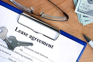 Clip board with lease agreement and keys on it with money, glasses, and a pen on a table