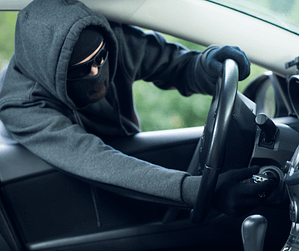 Person in hoodie, mask, and dark glasses leaning in car window and trying to start car