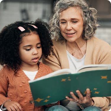 An older woman with grey curly hair sits close to a young girl with dark curly hair as they read an open green picture book together.