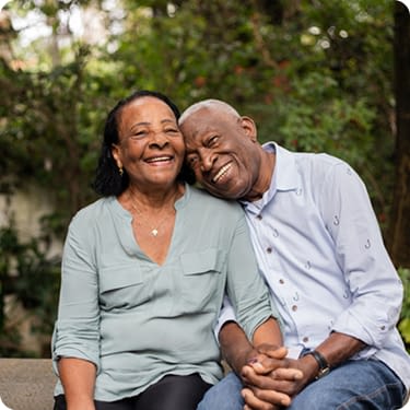 A smiling senior Black couple sitting close together outdoors, with the man resting his head affectionately against the woman’s shoulder as they hold hands. A smiling senior Black couple sitting close together outdoors, with the man resting his head affectionately against the woman's shoulder as they hold hands.