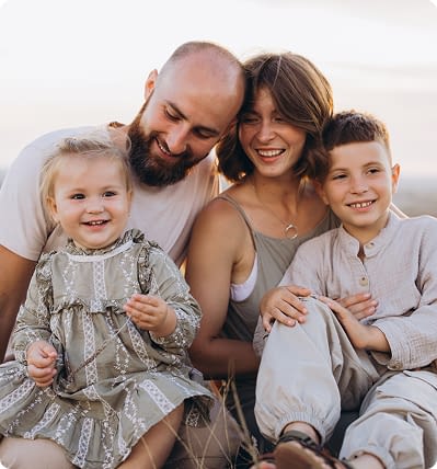 A close-up portrait of a family of four sitting together outdoors, including a mother, father, young son, and toddler daughter, all smiling gently.