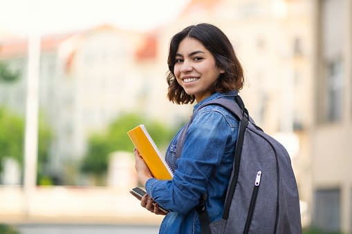 gettyimages-1473707800-170667a Smiling Young Arab Female Student With Workbooks And Backpack Standing Outdoors, Happy Middle Eastern Millennial Woman Posing Outside At University Campus, Turning At Camera, Copy Space