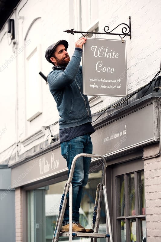 A man on a ladder fixing a name sign A man on a ladder fixing a painted name sign onto a bracket on a shopfront.