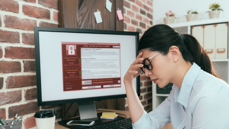 Cyber Insurance 7226 Female businesswoman sitting at desk with hand on forehead in frustration, hacked computer screen in background