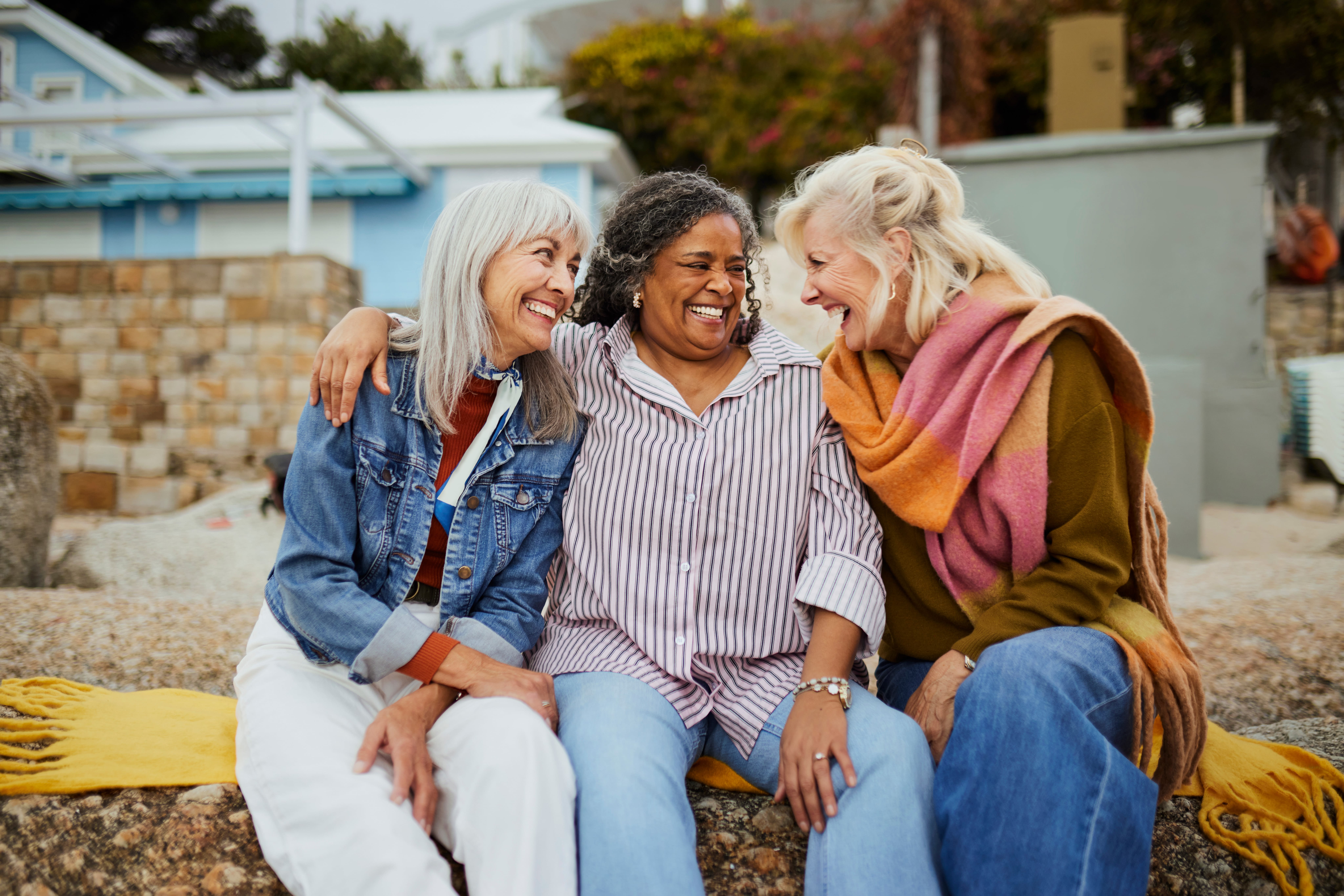 Three diverse senior women -- a silver-haired Caucasian, a Black woman, and a blonde Caucasian -- lean toward each other in a joyful shared laugh while sitting together at a coastal outdoor setting.